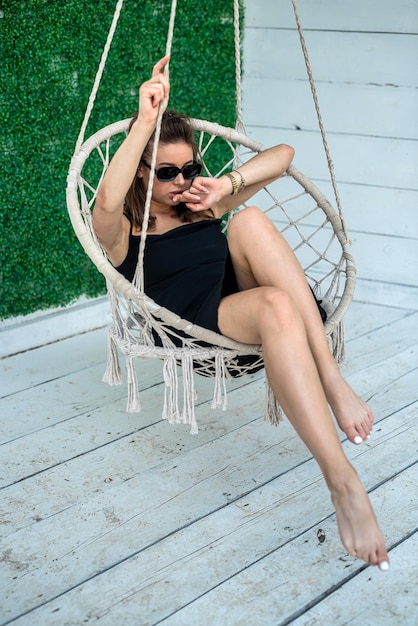 Hermosa modelo con un vestido negro y gafas de sol descansando en verano sentada en una mecedora