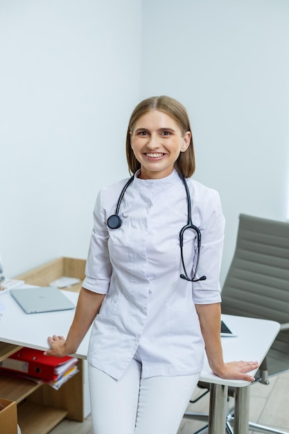 Una hermosa y moderna doctora con una bata médica blanca y un fonendoscopio está parada en la oficina mirando a la cámara Forma de aprendiz positiva Espacio libre para texto