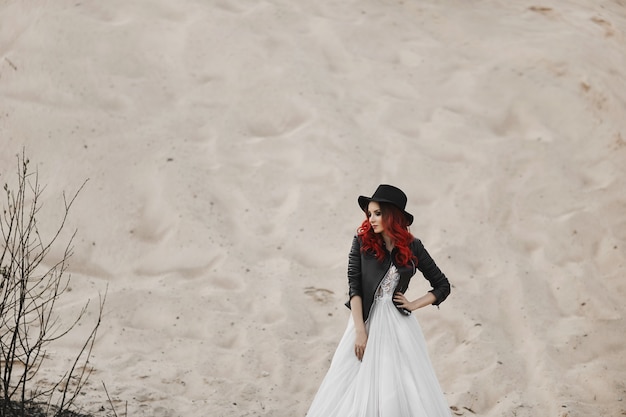 Hermosa y moderna modelo pelirroja con chaqueta de cuero, sombrero negro y elegante vestido de novia posando frente a una duna de arena