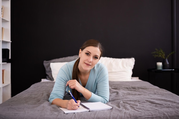 Una hermosa morena se acuesta en la cama de la habitación y toma notas en un cuaderno. Mantener un diario personal