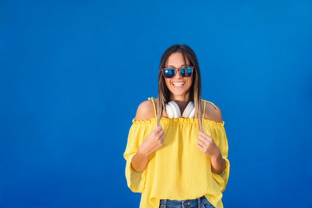 Hermosa morena en blusa amarilla, con gafas de sol y auriculares alrededor del cuello posando delante de la pared azul.