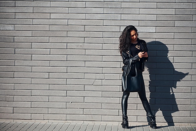 Hermosa morena con cabello rizado y ropa negra al aire libre cerca de la pared