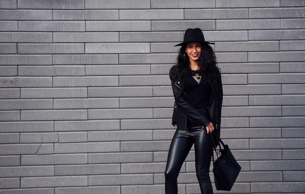 Hermosa morena con cabello rizado y ropa negra sosteniendo una bolsa al aire libre cerca de la pared