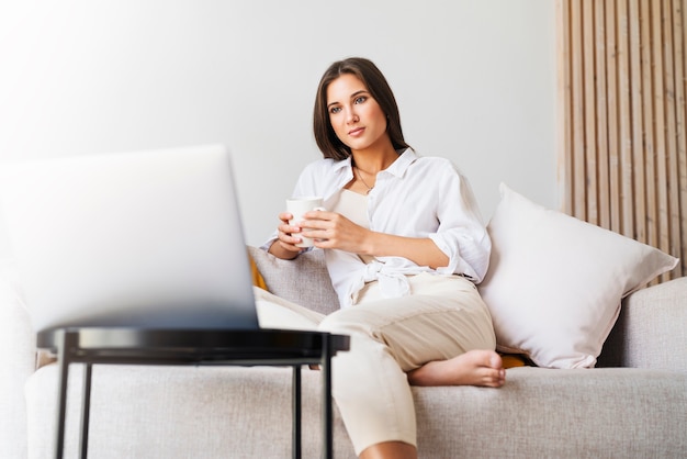 Hermosa morena con camisa blanca se sienta en el sofá, sosteniendo una taza blanca con café caliente en la mano.