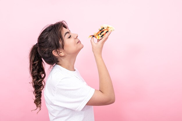 Hermosa morena con una camiseta blanca con un trozo de pizza sobre un fondo rosa, copie el espacio.