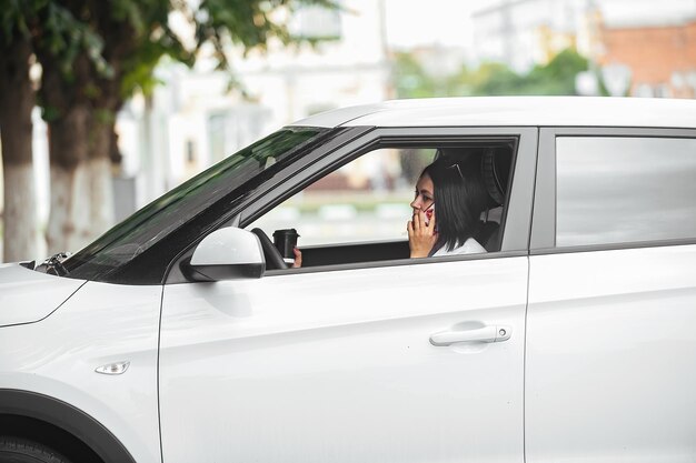 Hermosa morena conductora sentada en su auto con un vaso de café y hablando por teléfono