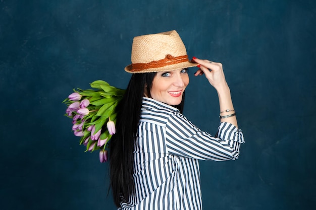 Hermosa morena de moda con sombrero de paja de verano y camisa a rayas con un ramo de tulipanes