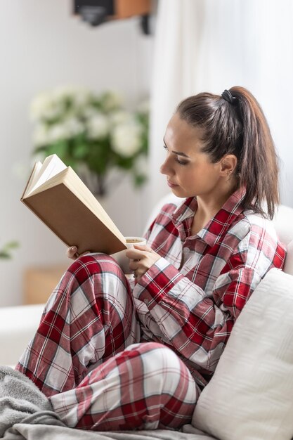 Hermosa morena en pijama está leyendo un libro sentada en el sofá de la sala de estar