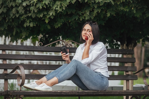 Hermosa morena sentada en un banco con un vaso de bebida en el parque y hablando por teléfono