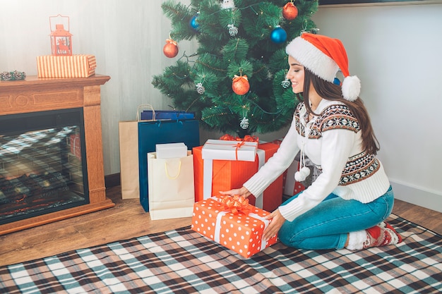 Hermosa morena sentada en posición de escuadrón en el árbol de Navidad con regalos y tiene caja roja