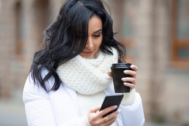 Hermosa morena con un teléfono inteligente y un café en la calle de una ciudad europea en la temporada de invierno