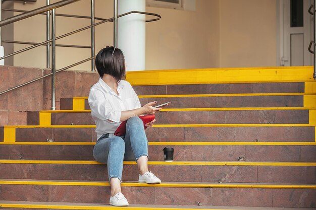 una hermosa morena con un teléfono y una tableta se sienta en los escalones frente a la puerta y espera