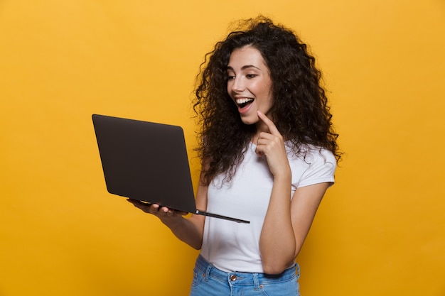 Hermosa mujer de 20 años vistiendo ropa casual sonriendo mientras sostiene portátil negro aislado en amarillo