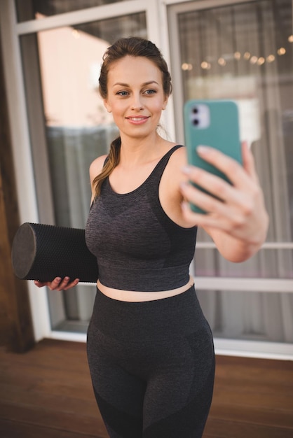 Hermosa mujer de 25 a 30 años se prepara para hacer ejercicio en casa haciendo selfie sosteniendo un teléfono inteligente en el interior. Estilo de vida saludable. La chica feliz hace ejercicio en línea y tiene videollamadas.