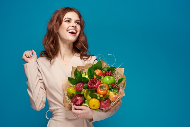 Hermosa mujer abrigo beige ramo de frutas en manos fondo azul