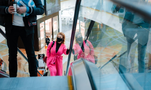 Hermosa mujer abrigo brillante paseos en escaleras mecánicas centro comercial máscara protectora negra en su cara contra el aire contaminado por virus