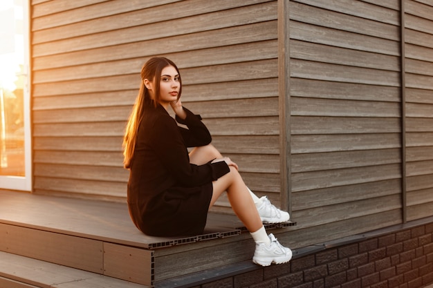 Hermosa mujer con un abrigo de moda con zapatos blancos sentado cerca de una casa de madera vintage al atardecer