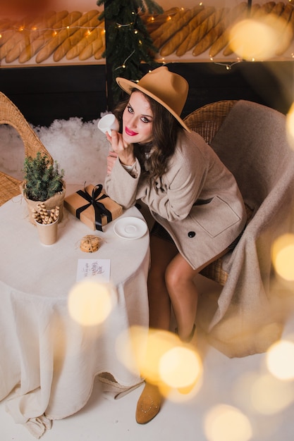 Una hermosa mujer con un abrigo y un sombrero se sienta en una cafetería con un regalo en la mano concepto de Navidad