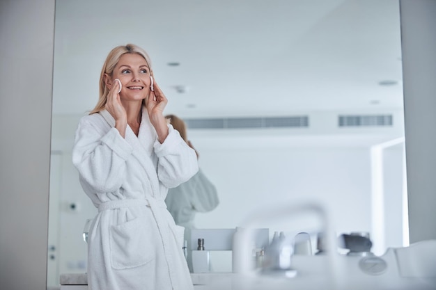 Hermosa mujer adulta en albornoz de felpa blanca limpiando la cara de la piel en el baño