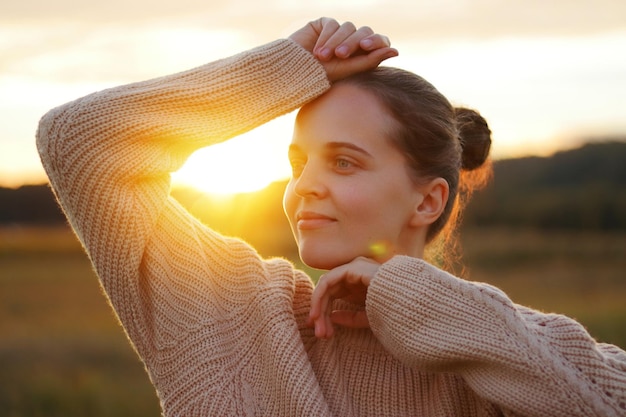 Hermosa mujer adulta joven con suéter beige de pie con los brazos levantados y mirando hacia otro lado con expresión facial de ensueño posando en la naturaleza al aire libre