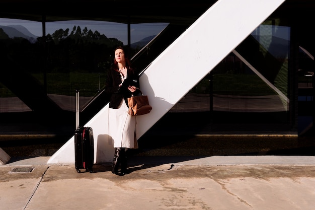 Foto hermosa mujer adulta joven en viaje de negocios pensativo en la calle revisando el correo electrónico por zona de negocios o parque industrial con maleta. concepto de viaje de negocios