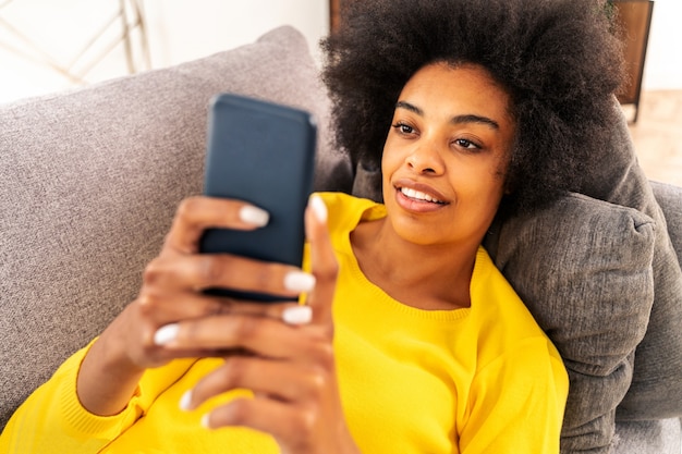 Hermosa mujer adulta negra en casa viendo videos en línea en la aplicación de red social con teléfono inteligente - Chica bastante negra trabajando en línea remota en el teléfono móvil