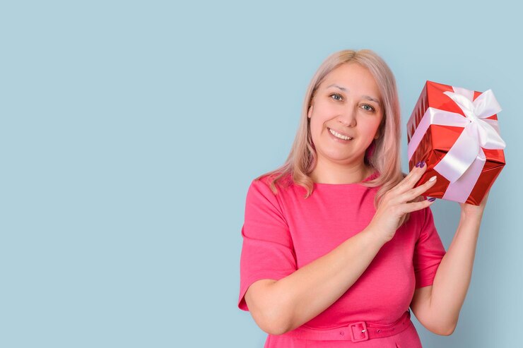Una hermosa mujer adulta rubia sacude una caja de regalo junto a su oreja tratando de adivinar qué hay dentro de pie sobre un fondo azul, espacio de copia
