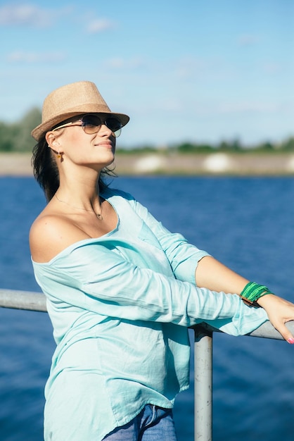 Hermosa mujer adulta con sombrero y gafas de sol camina en el parque junto al lago en un día soleado de verano