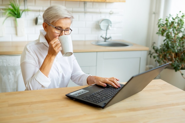 Hermosa mujer adulta en vasos en la mesa con un portátil en casa con una taza