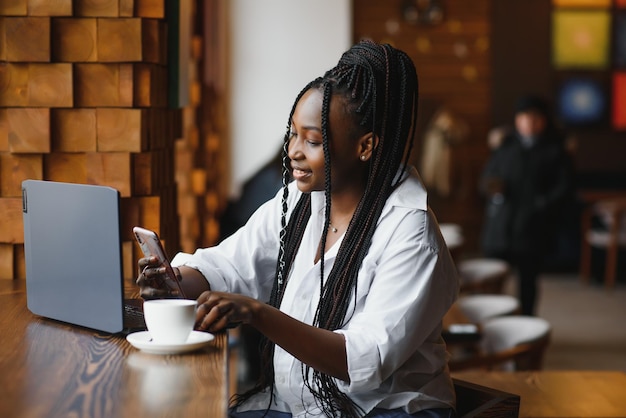 Hermosa mujer africana con cabello largo y oscuro está escribiendo mensajes en un teléfono inteligente mientras está sentada junto a la ventana en una luz suave. Una joven mujer de negocios está usando una computadora portátil y un teléfono móvil para el trabajo.