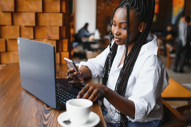 Hermosa mujer africana con cabello largo y oscuro está escribiendo mensajes en un teléfono inteligente mientras está sentada junto a la ventana en una luz suave. Una joven mujer de negocios está usando una computadora portátil y un teléfono móvil para el trabajo.