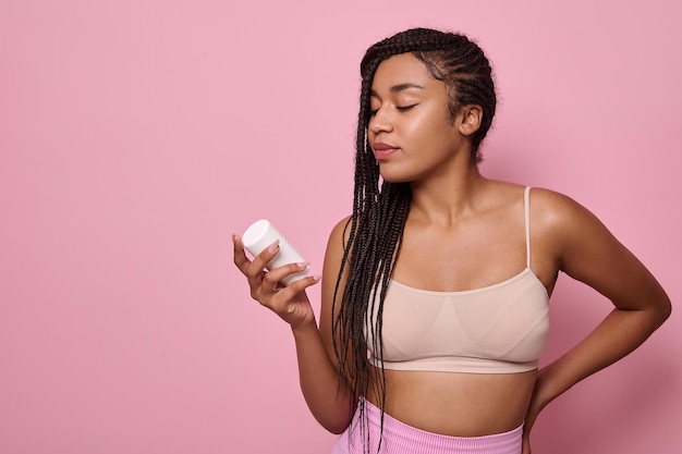 Hermosa mujer africana con elegantes rastas afro en ropa interior sosteniendo una botella con producto cosmético de belleza, aislado sobre fondo rosa con espacio de copia para publicidad