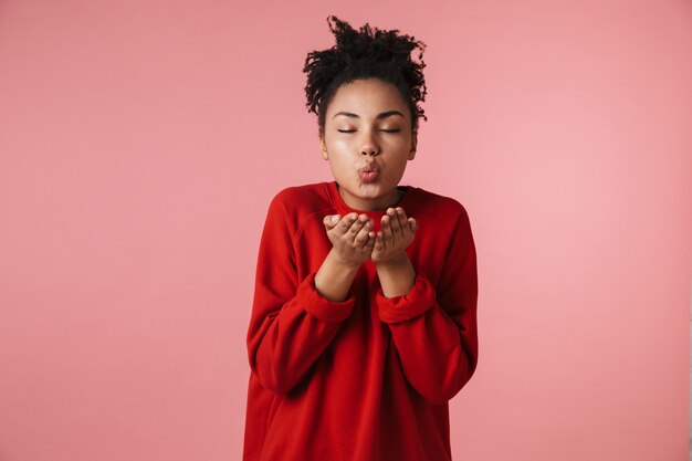 Hermosa mujer africana emocionada feliz joven asombrosa posando aislada sobre pared rosa soplando besos.