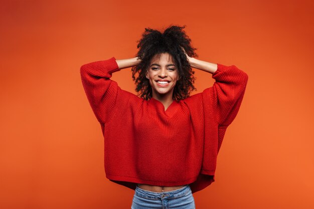 Hermosa mujer africana joven alegre vistiendo suéter de pie islolated sobre pared roja, posando, jugando con el pelo