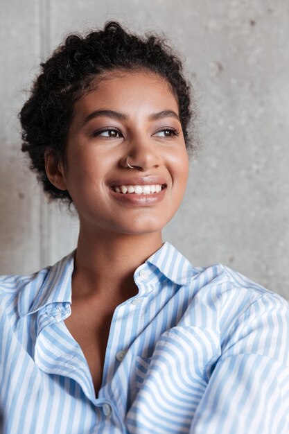 Hermosa mujer africana joven sonriente con ropa casual y gafas, apoyado en una pared en el interior, posando mientras está de pie