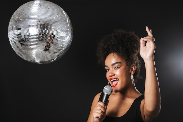 Hermosa mujer africana de pie con bola de discoteca plateada aislada sobre espacio negro, sosteniendo el micrófono