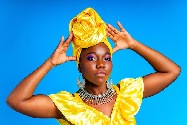 Hermosa mujer africana en turbante de seda amarilla en la cabeza y vestido con plata sin cuello y aretes piercing en la nariz en el fondo azul del estudio