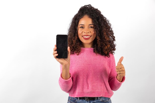 Hermosa mujer afro sonriendo y con el pulgar hacia arriba sosteniendo un teléfono inteligente con la pantalla en blanco para texto