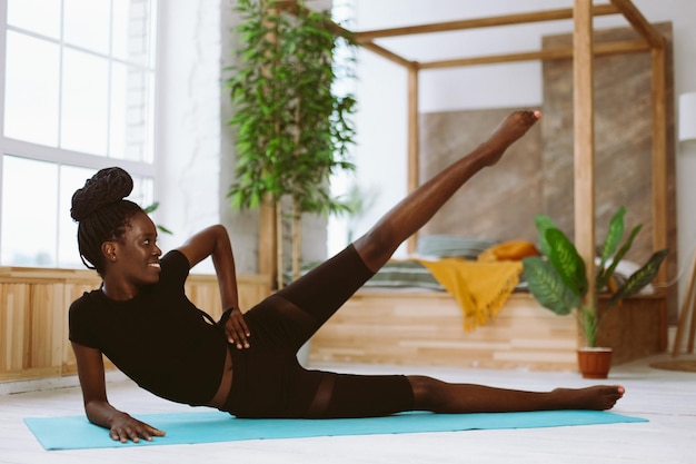 Hermosa mujer afroamericana atlética acostada de lado y levantando la pierna hasta el entrenamiento de enfoque selectivo en la alfombra del gimnasio en un estudio fotográfico decorado Curso deportivo técnica de ejercicio fitness mantener el cuerpo en forma