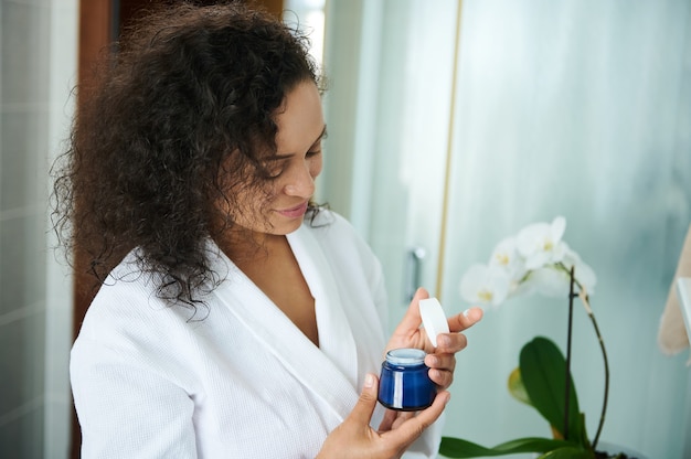 Foto hermosa mujer afroamericana en una bata de baño blanca en el baño abre un frasco de crema hidratante