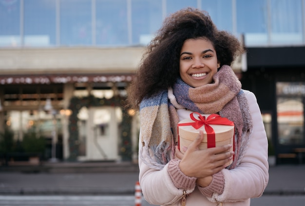 Hermosa mujer afroamericana con caja de regalo, de pie en la calle urbana, sonriendo