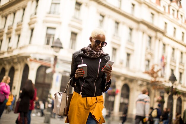 Hermosa mujer afroamericana elegante con café y el uso de un teléfono inteligente