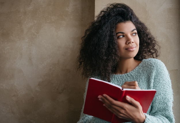 Hermosa mujer afroamericana con libro rojo, mirando por la ventana