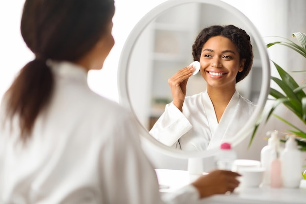 Hermosa mujer afroamericana limpiando la piel con almohadilla de algodón cerca del espejo