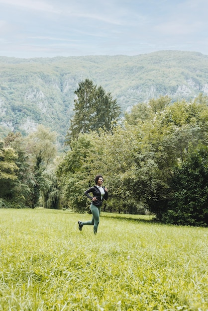 Hermosa mujer afroamericana madura sonriente está trotando a través de la hierba verde cerca del paisaje montañoso.