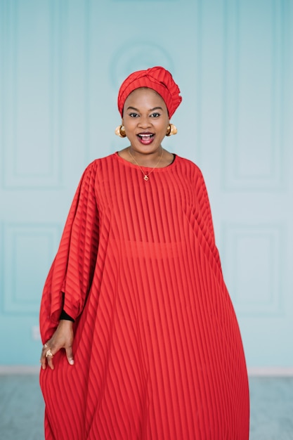 Hermosa mujer afroamericana positiva vestida con ropa étnica roja y pañuelo en la cabeza, caminando en cámara con cara feliz sonriendo. Foto de estudio de señora bonita sobre fondo azul.