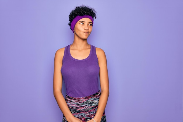 Hermosa mujer afroamericana con ropa deportiva informal sobre fondo púrpura sonriendo mirando hacia un lado y mirando hacia otro lado pensando