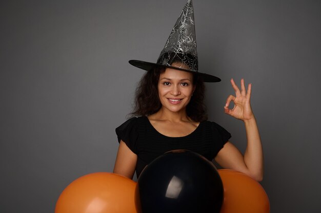 Hermosa mujer afroamericana sonriente con sombrero de mago, vestida con un disfraz de carnaval de brujas para la fiesta de Halloween, muestra el signo de OK, posa sobre un fondo gris con globos negros, espacio de copia