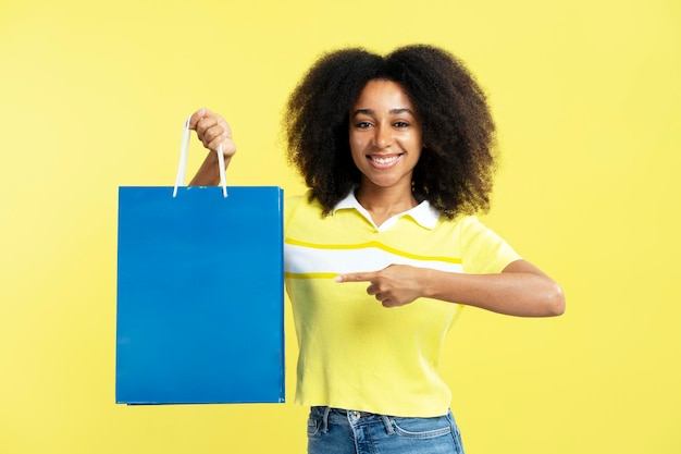 Hermosa mujer afroamericana sonriente sosteniendo una bolsa de compras azul aislada sobre fondo amarillo