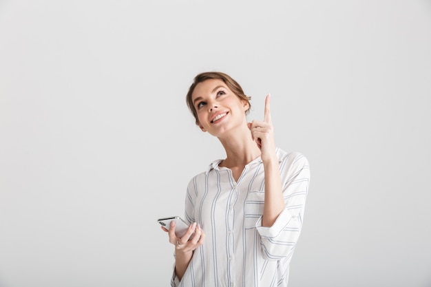 Hermosa mujer alegre apuntando con el dedo hacia arriba y usando el teléfono celular aislado sobre fondo blanco.